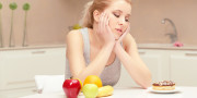 It is hard to make a choice. Young woman sitting at her table in the kitchen and trying to choose eating either banana, apple and orange or sweet donut