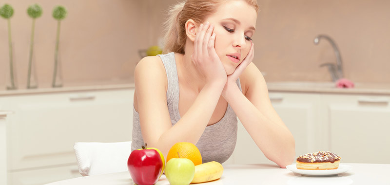 It is hard to make a choice. Young woman sitting at her table in the kitchen and trying to choose eating either banana, apple and orange or sweet donut