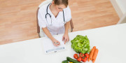 High Angle View Of Female Dietician Writing Prescription With Vegetables On Desk