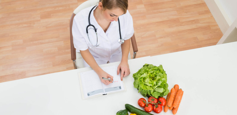 High Angle View Of Female Dietician Writing Prescription With Vegetables On Desk